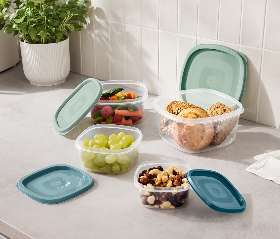 Mutfak tezgahında üzüm, doğranmış sebzeler, kuruyemiş ve kurabiyelerle dolu, mavi-yeşil kapaklı dört adet şeffaf kap bulunmaktadır.