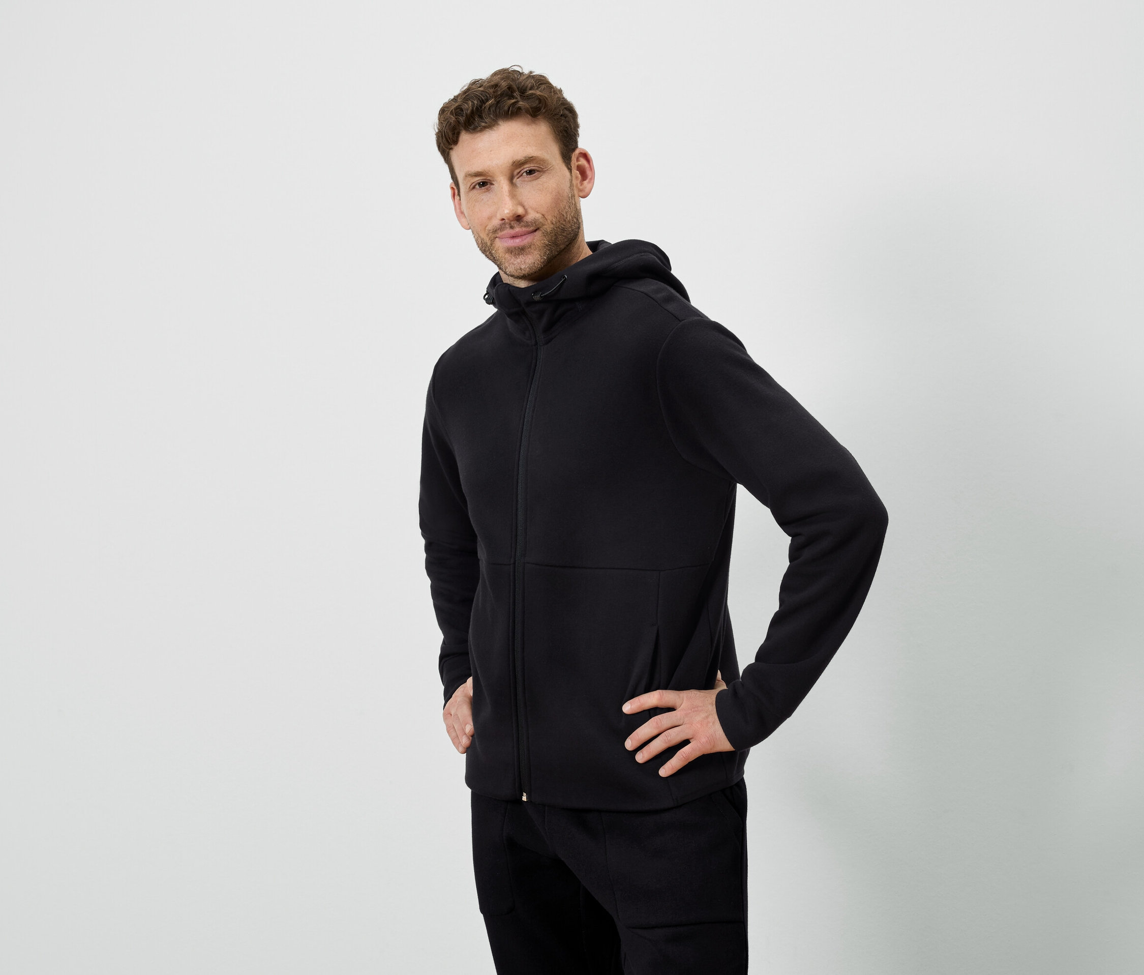 Beyaz bir arka planda poz veren bir adam. Siyah kapüşonlu bir sweatshirt ve siyah pantolon giyiyor.