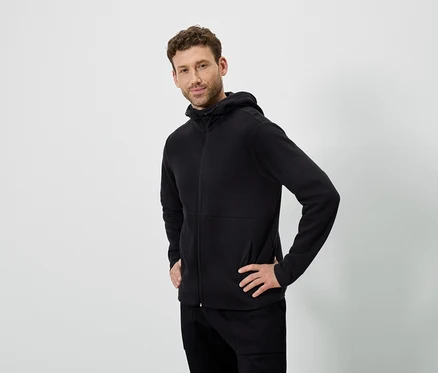 Beyaz bir arka planda poz veren bir adam. Siyah kapüşonlu bir sweatshirt ve siyah pantolon giyiyor.