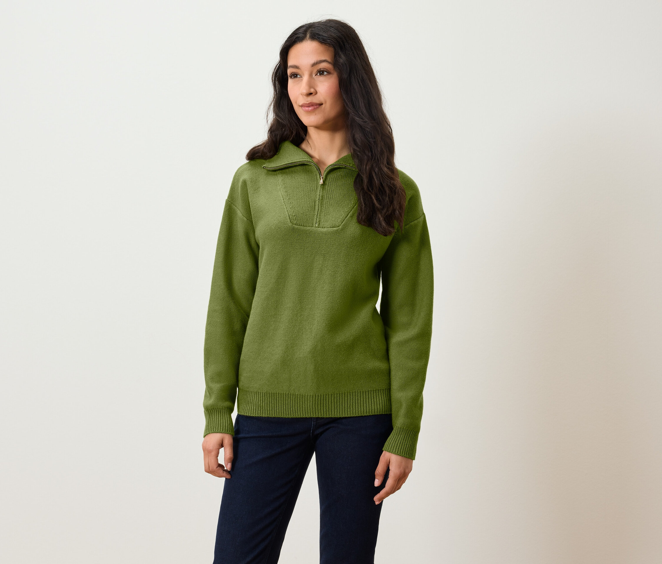 Bir kadın ayakta duruyor, üzerinde kısa fermuarlı zeytin yeşili bir kazak ve lacivert pantolon var.