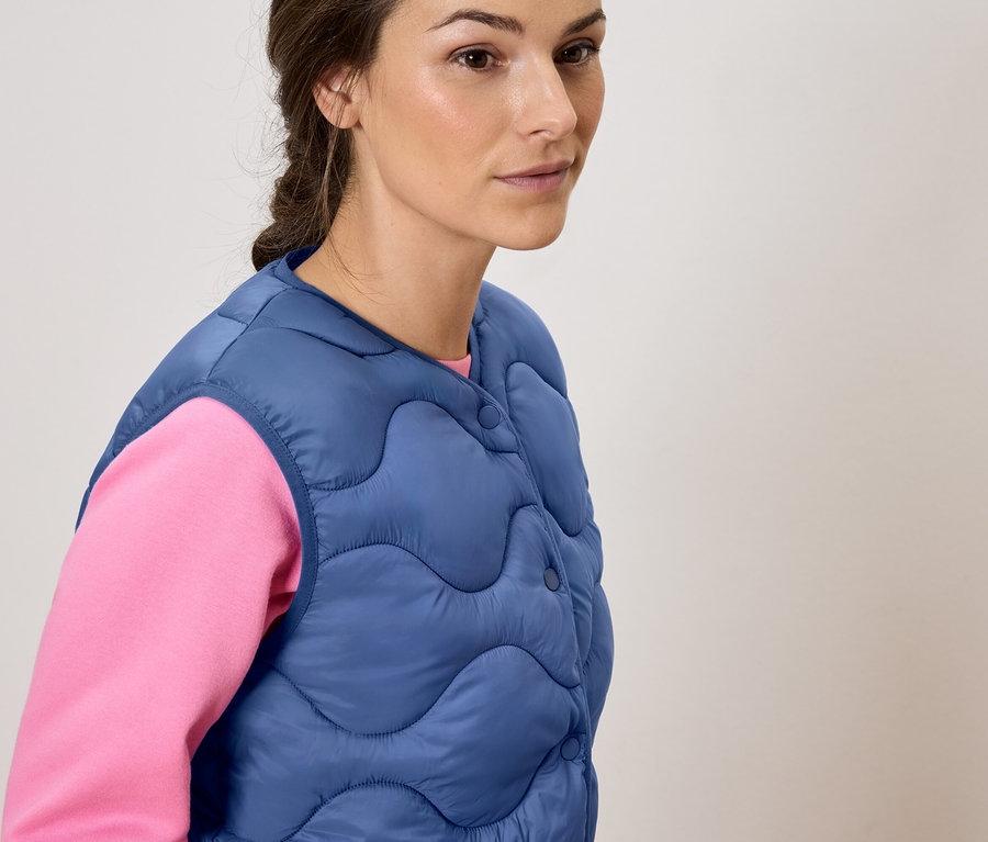 Kadın pembe bir bluz üzerine mavi kapitone bir yelek giyiyor.