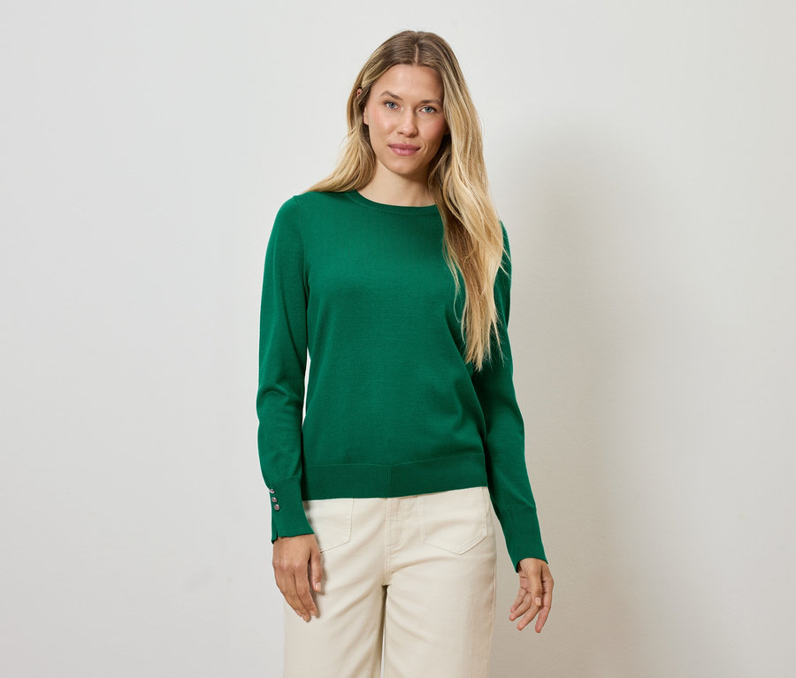 Sarışın bir kadın, yeşil bir kazak ve krem rengi pantolonla beyaz bir arka plan önünde duruyor.