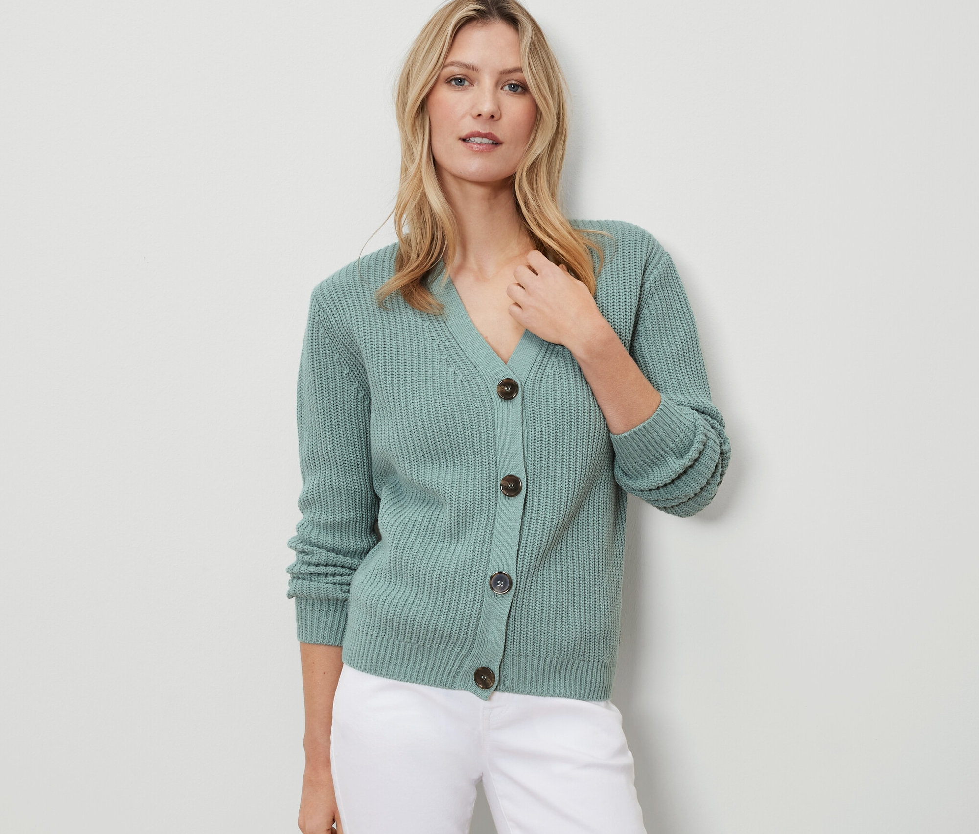 Kadın adaçayı rengi, kalın örgü bir hırka ve beyaz pantolon giyiyor.