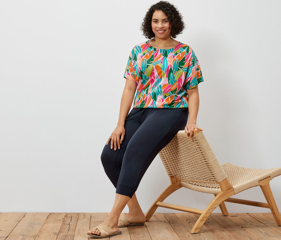 Bir kadın hasır bir sandalyede oturuyor, lacivert 3/4 jersey pantolon ve tamamı baskılı düşük omuzlu tişört giyiyor.