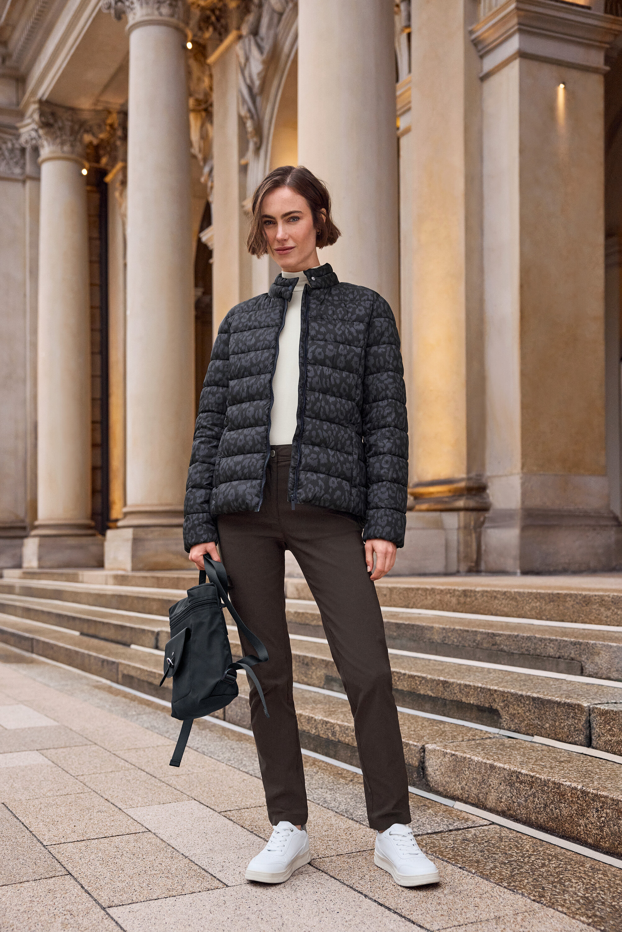 Kadın merdivenlerin önünde duruyor, leopar desenli bir şişme mont, kahverengi pantolon ve beyaz Tom Tailor PU spor ayakkabı giyiyor.