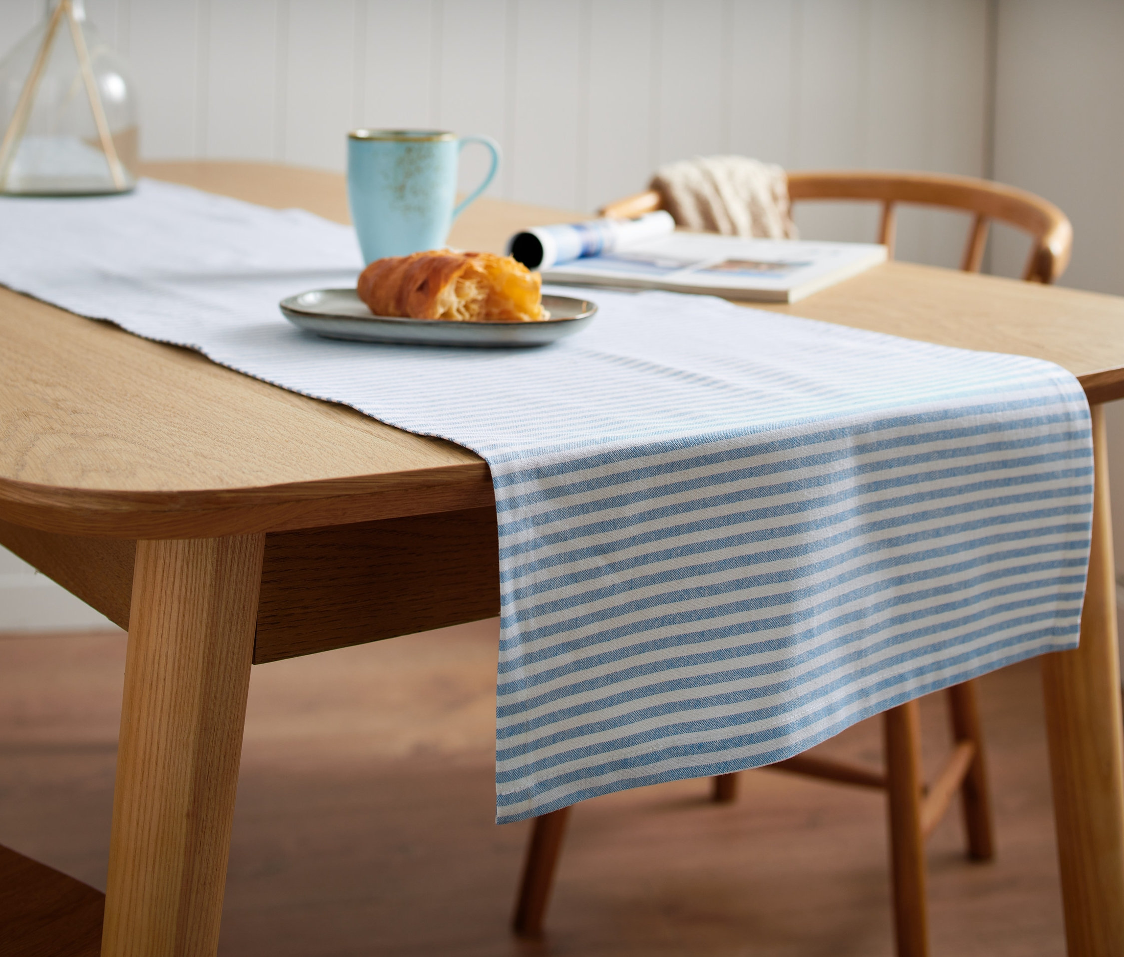 Ahşap bir masa üzerinde mavi beyaz çizgili runner masa örtüsü bulunuyor. Üzerinde hamur işi olan bir tabak ve mavi bir fincan var.