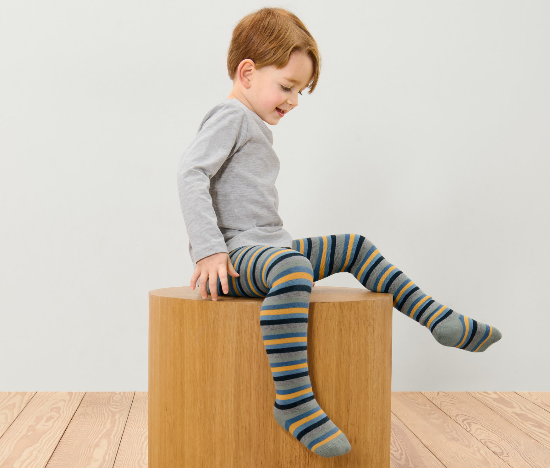 Bir çocuk ahşap bir taburede oturuyor ve üzerinde 2 ADET MAVİ VE ÇOK RENKLİ ÇOCUK TERMAL KÜLOTLU ÇORAP var.