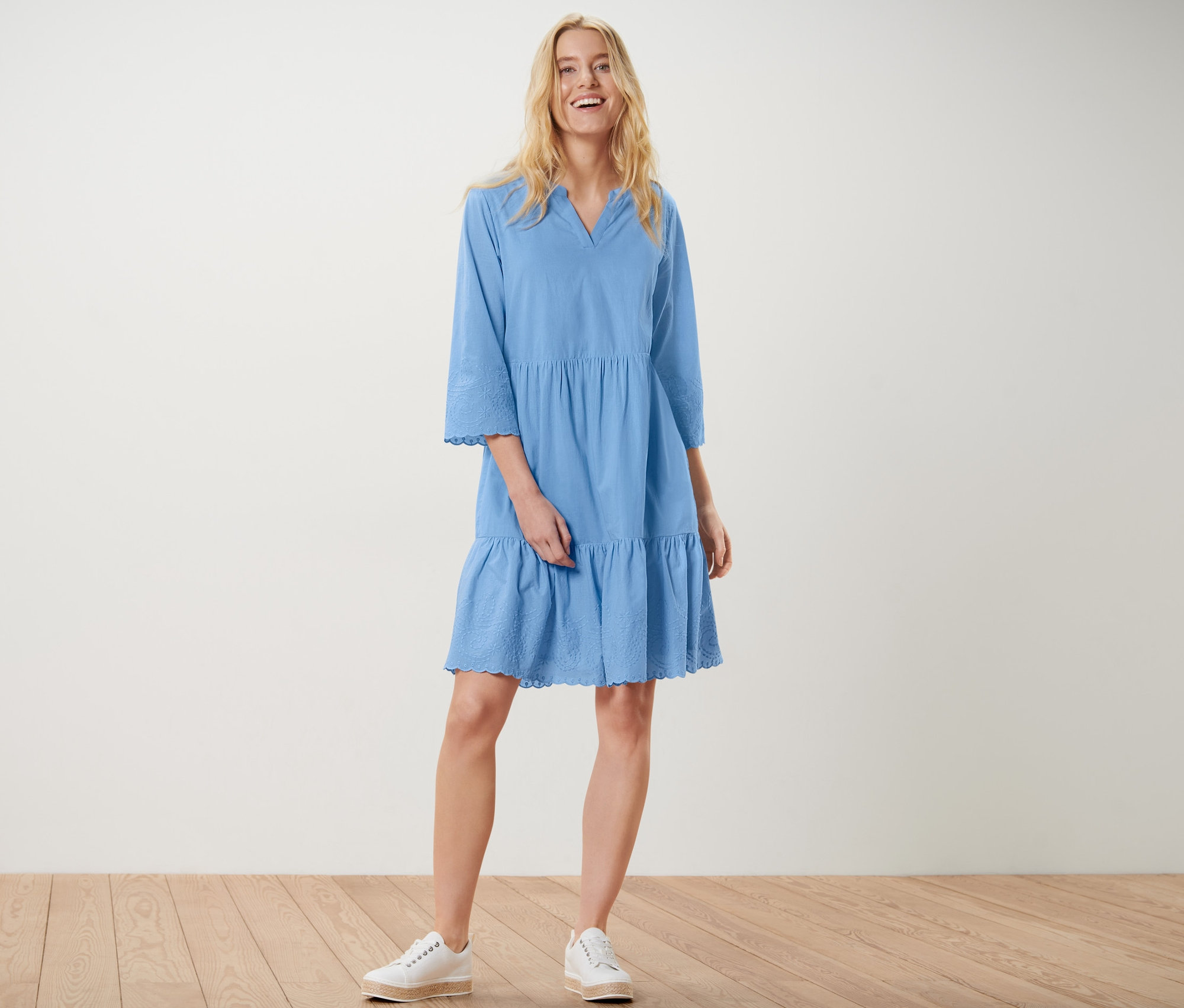 Sarışın bir kadın ayakta duruyor ve gülümsüyor, nakışlı mavi dokuma elbise ve beyaz spor ayakkabı giyiyor.