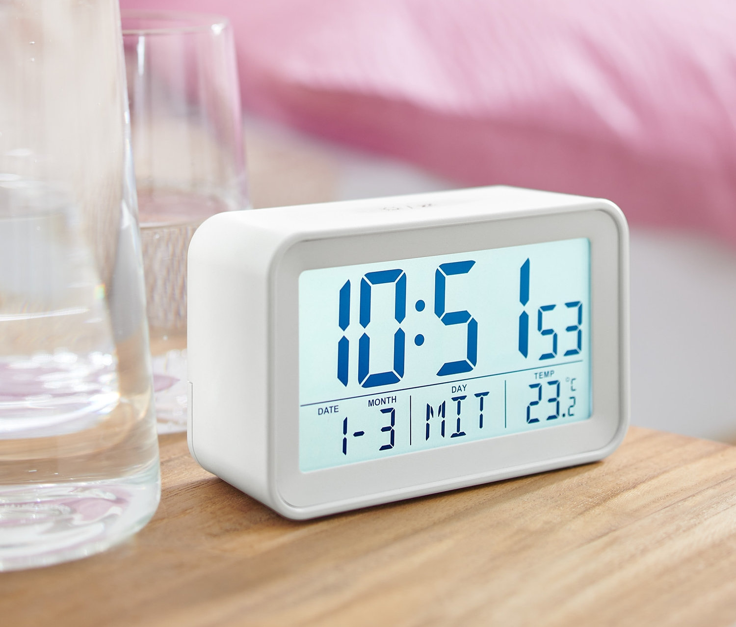 Beyaz bir dijital LCD çalar saat, bir bardak suyun yanında bir komodinin üzerinde duruyor.