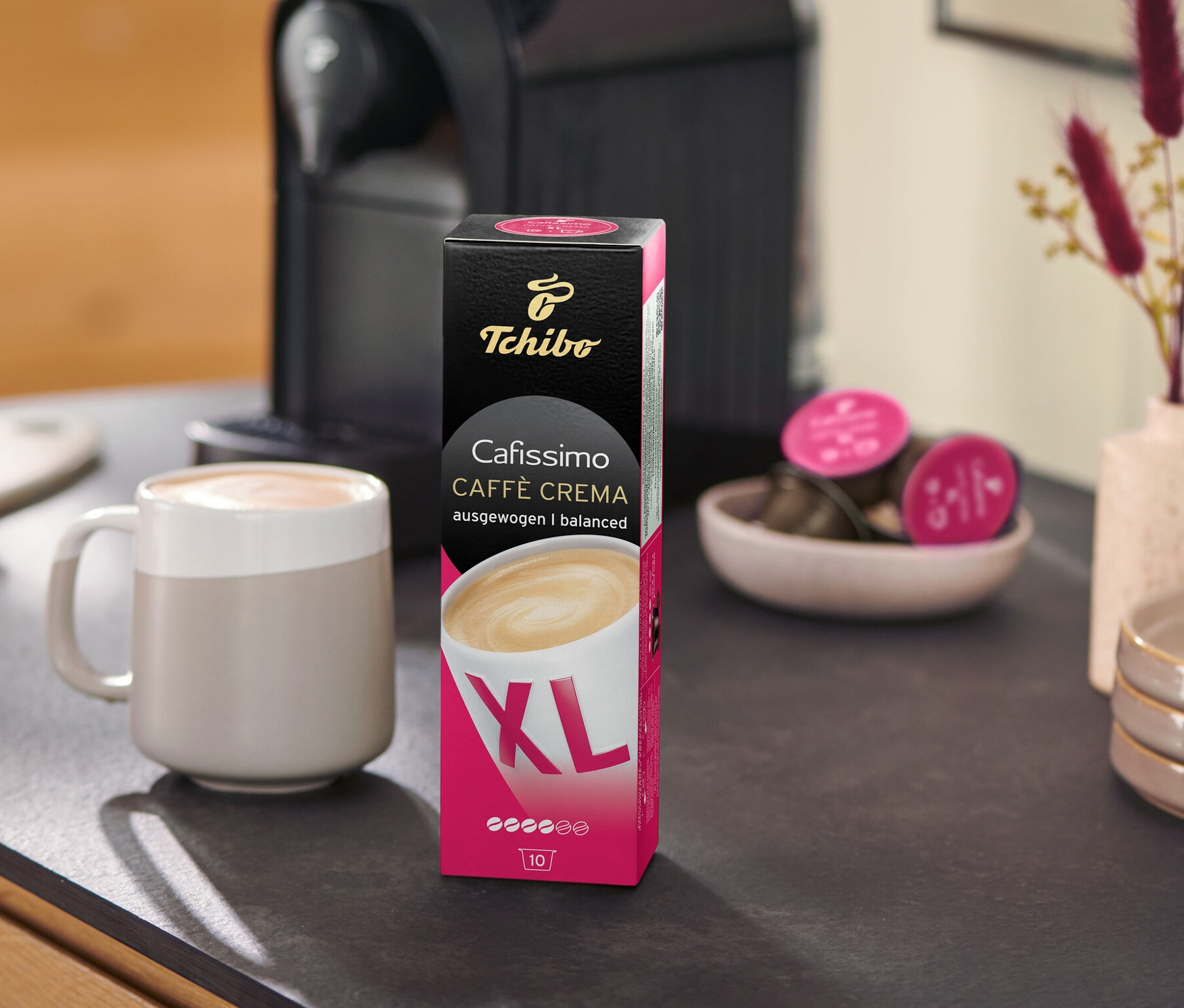 Tezgahta bir kahve makinesi, bir Caffè Crema Wake Up XL – 80 Kapsül paketi ve bir fincan bulunmaktadır.