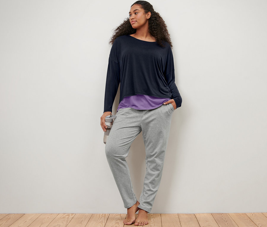 Duvara yaslanmış bir kadın, gri eşofman altı ve mavi-lila 2'si 1 arada spor tişörtü giyiyor.