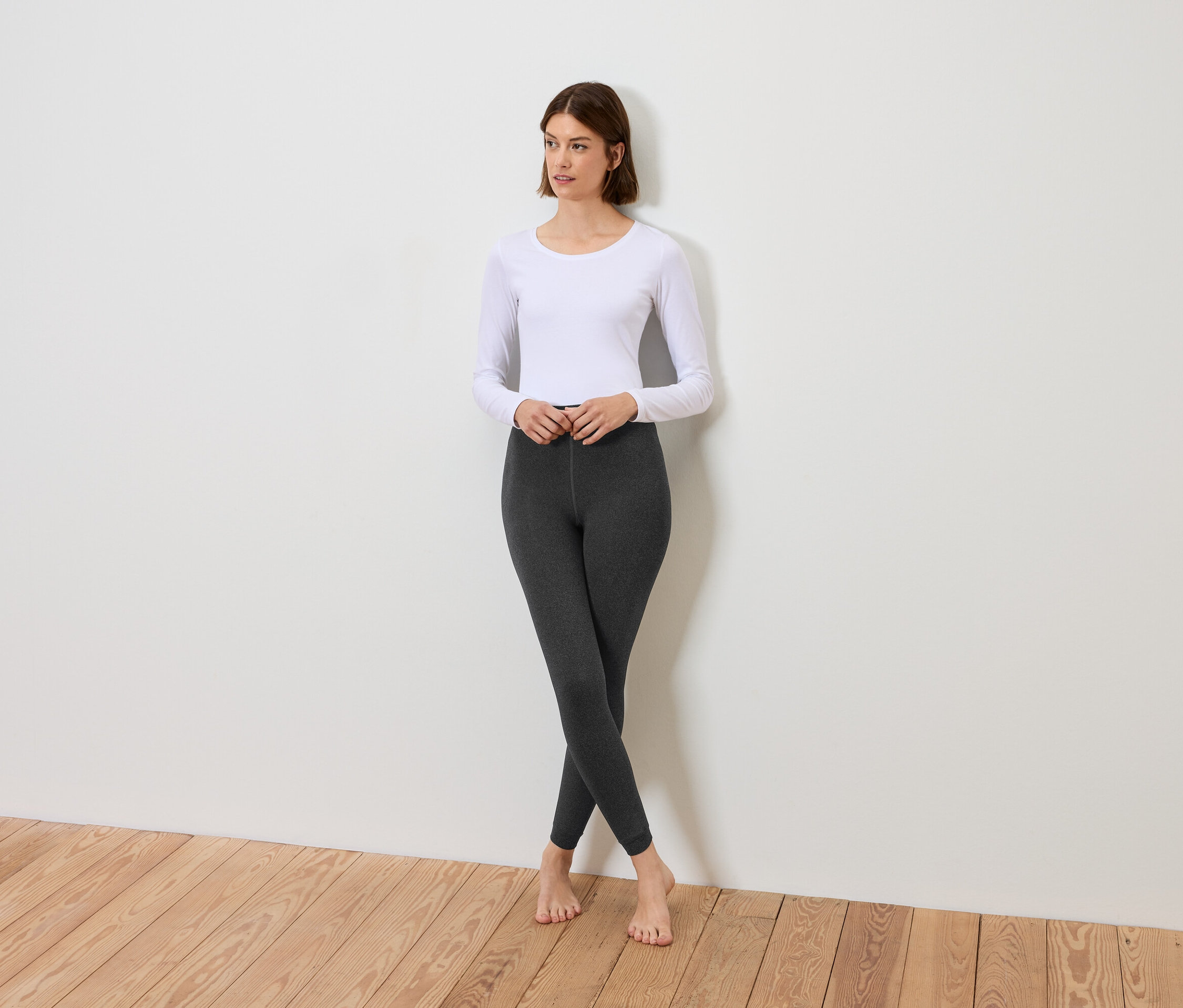 Bir kadın beyaz uzun kollu tişört ve gri tayt giyerek çapraz bacaklı duruyor.