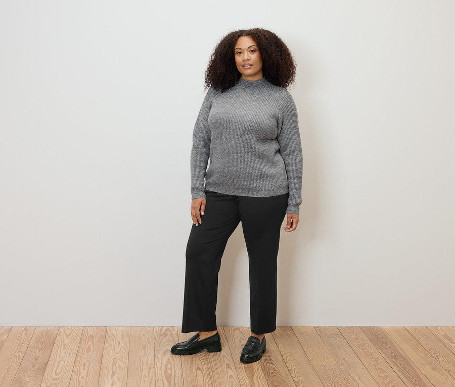 Kadın ayakta duruyor, gri boğazlı, kalın örgü bir kazak ve rahat kesim jarse bir pantolon giyiyor.