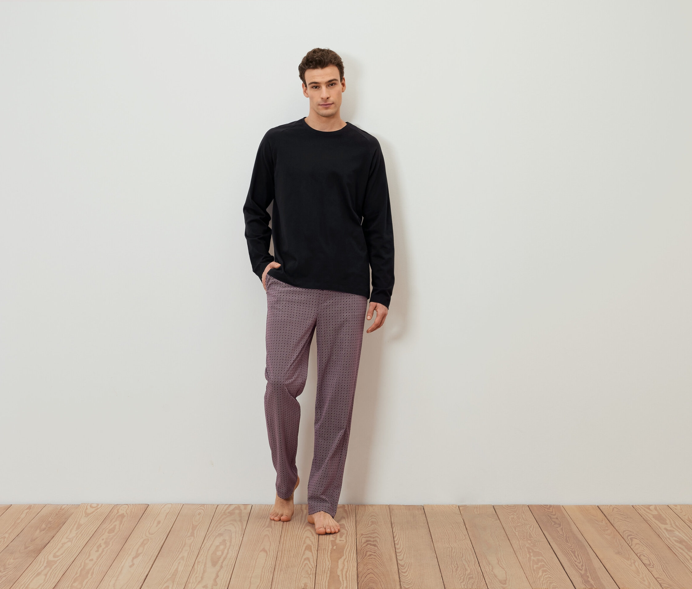 Adam beyaz bir duvarın önünde yalın ayak duruyor, üzerinde siyah uzun kollu bir tişört ve desenli bir pantolon var.