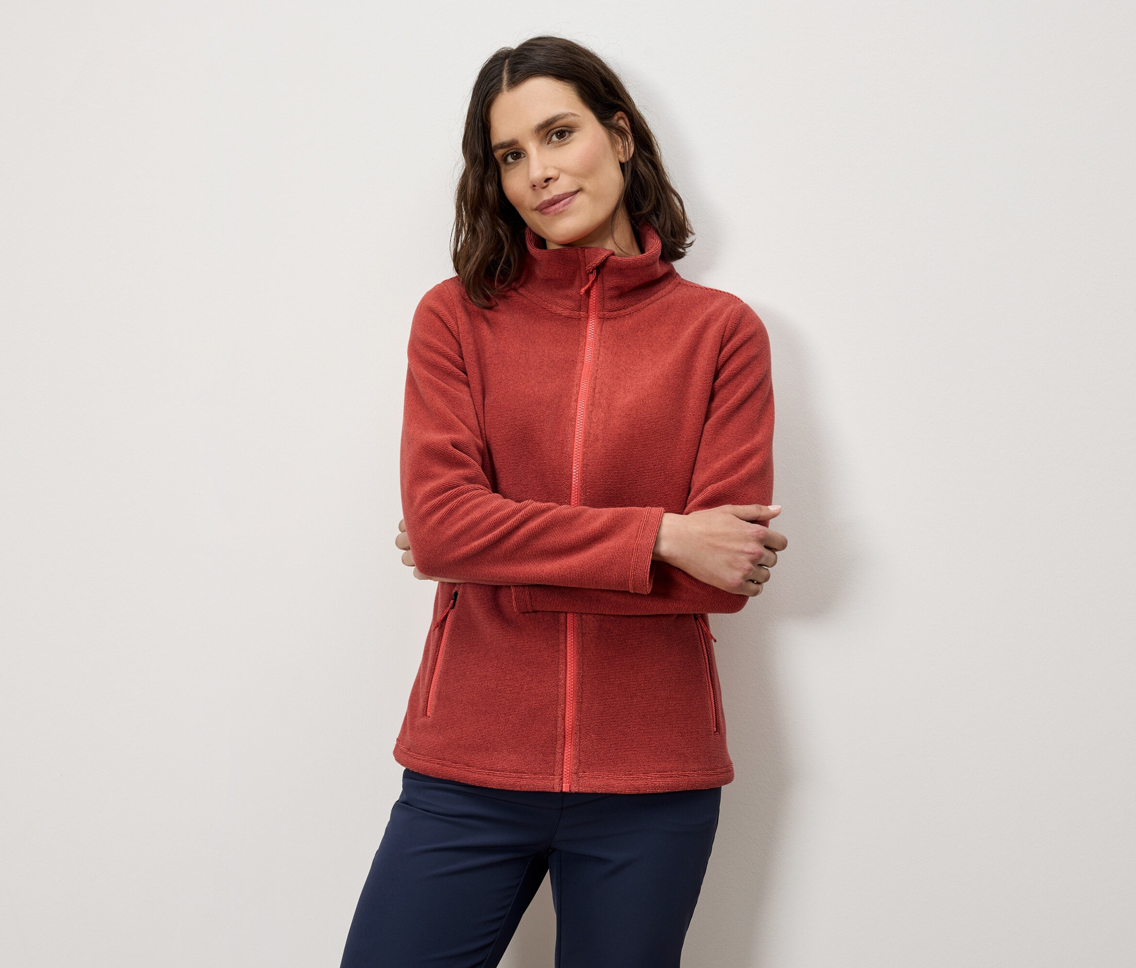 Kadın turuncu bir polar ceket ve lacivert pantolonla kolları kavuşturarak poz veriyor.