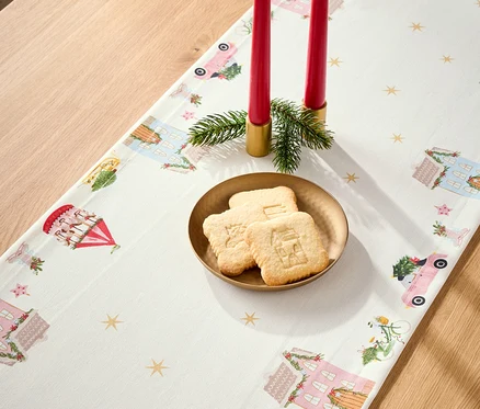 Masanın üzerinde ev, araba ve atlıkarınca motifleriyle süslenmiş bir Noel masa örtüsü, kırmızı mumlar ve bir tabak Noel kurabiyesi bulunmaktadır.