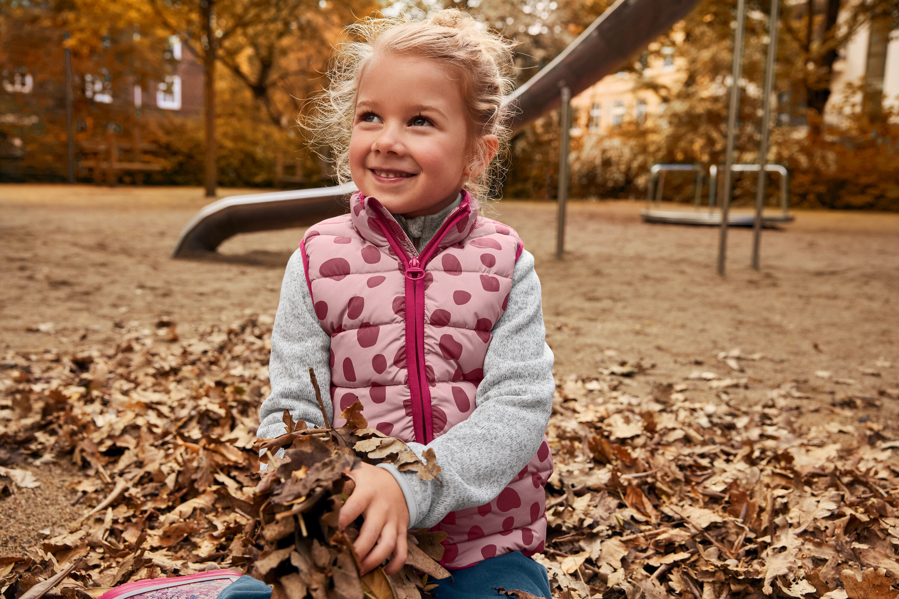Gülümseyen bir kız çocuğu oyun alanında bir yaprak yığınında oturuyor ve pembe puantiyeli bir yelek giyiyor.