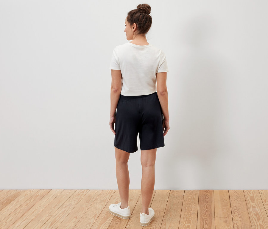 Bir kadın kameraya arkası dönük duruyor ve siyah jarse şort giyiyor.