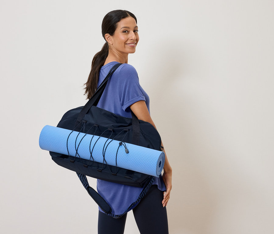 Kadın omzunda siyah bir spor çantası içinde mavi bir yoga matı taşıyor ve omzunun üzerinden bakıyor.