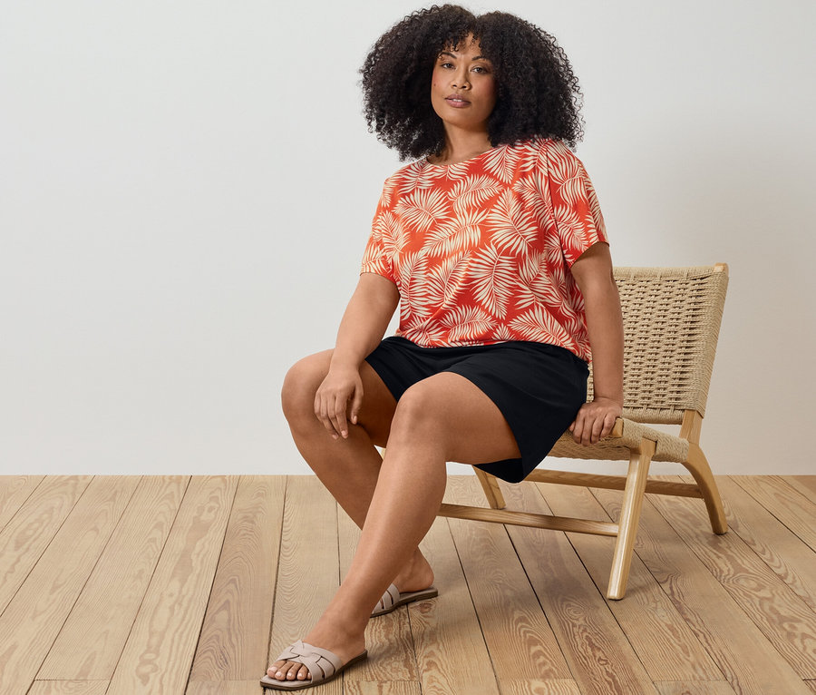 Bir kadın sandalyede oturuyor, üzerinde baskılı bir bluz ve siyah jersey şort var.