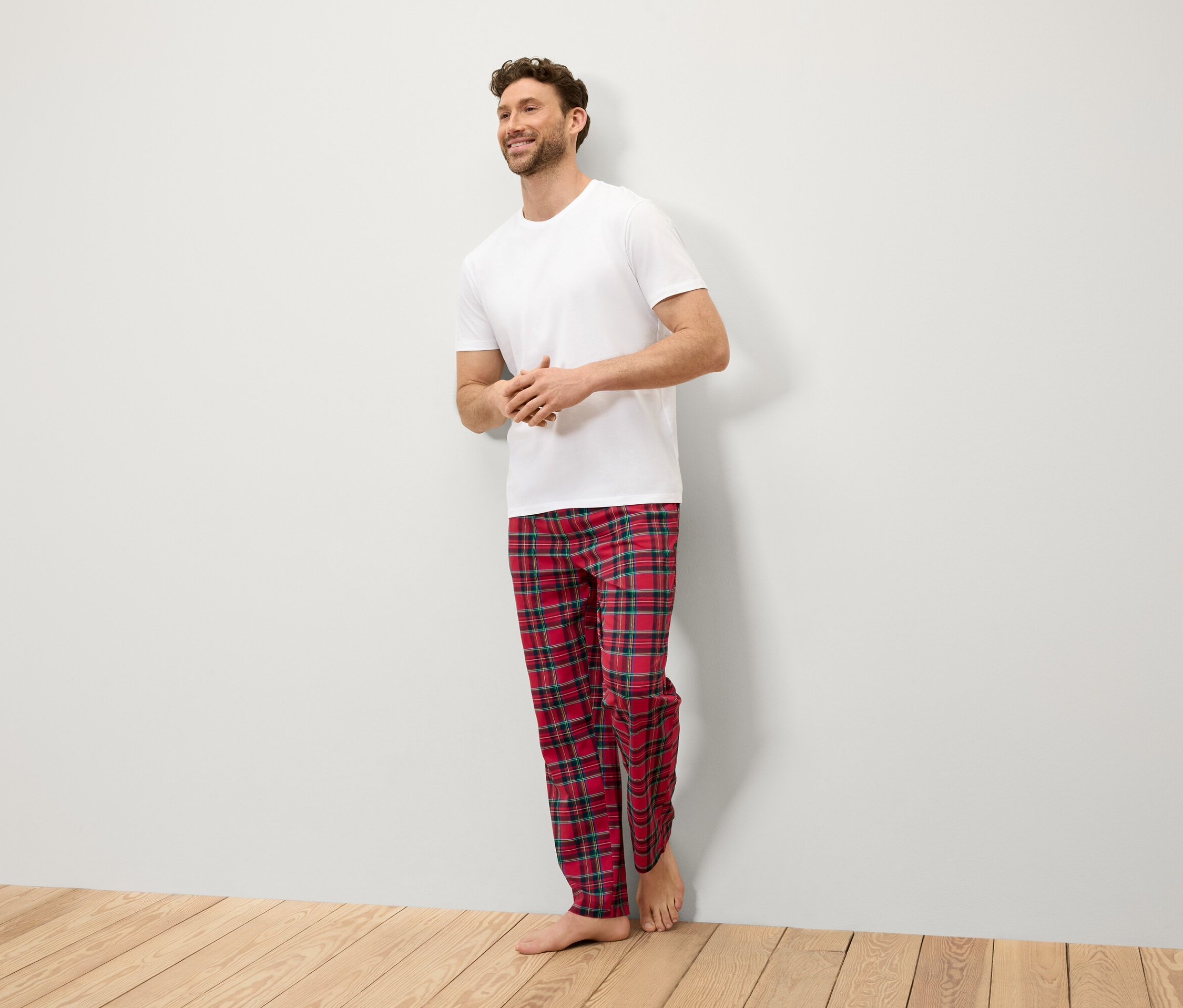 Gülümseyen bir adam duvara yaslanmış, beyaz bir tişört ve kırmızı kareli bir pantolon giyiyor.