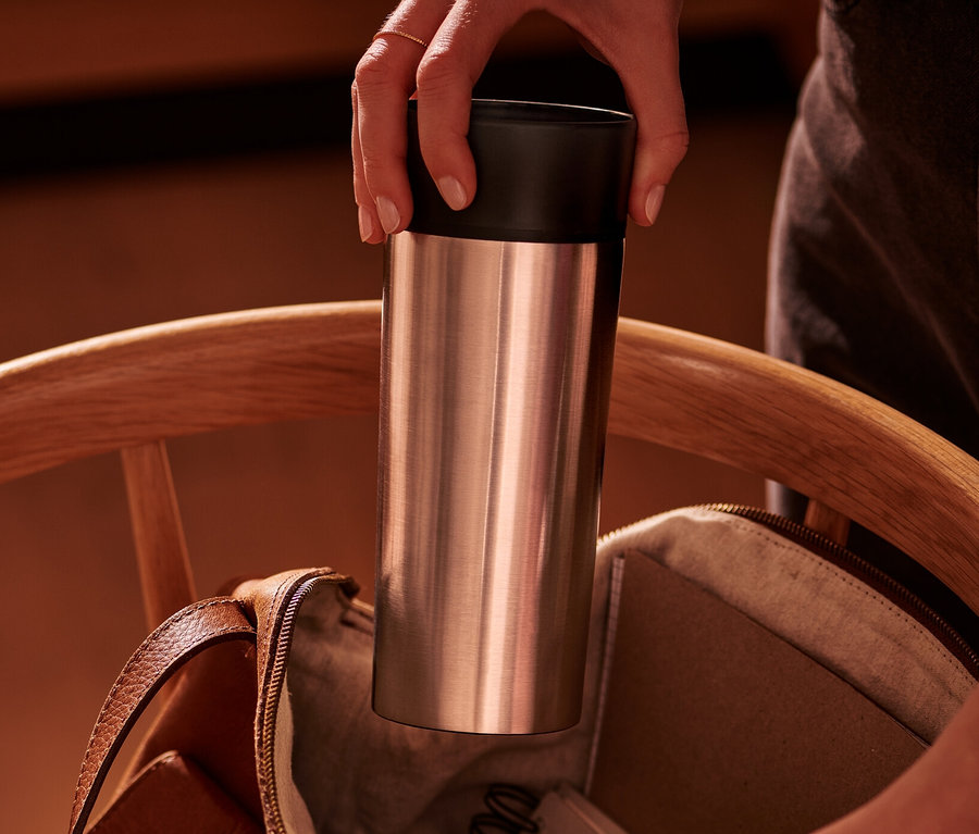 Bir el, paslanmaz çelik Coffee-to-go Kupası'nı ahşap sandalyedeki açık kahverengi deri çantaya yerleştiriyor.
