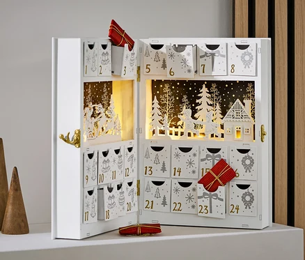Açık, beyaz renkli, süslemeli ve hediyeli bir advent takvimi, üç adet ahşap koninin yanında duruyor.
