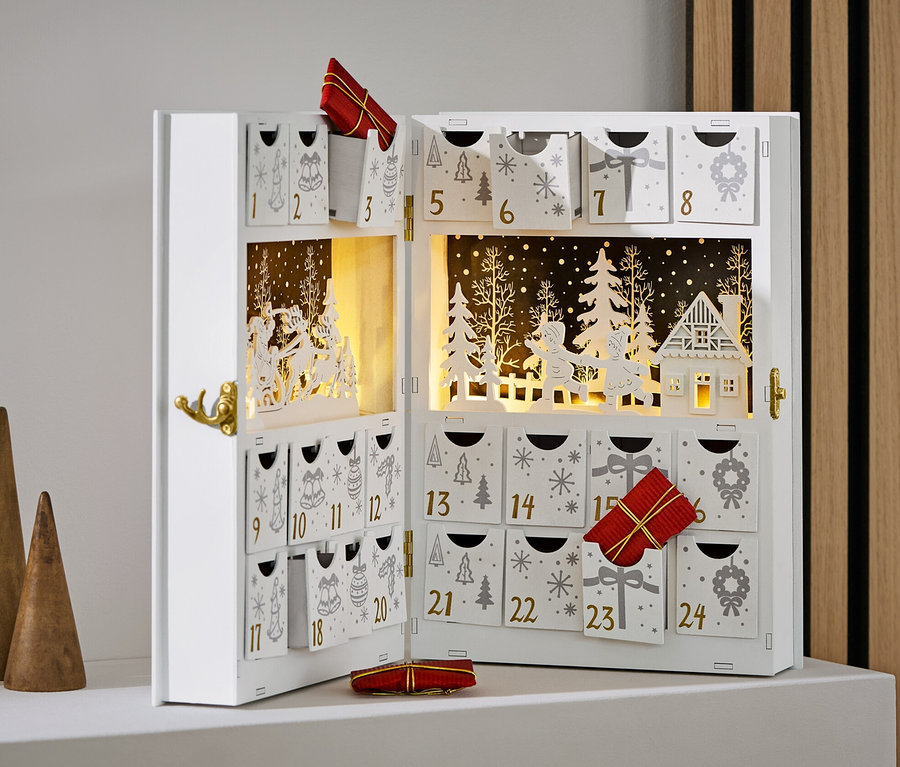 Açık, beyaz renkli, süslemeli ve hediyeli bir advent takvimi, üç adet ahşap koninin yanında duruyor.