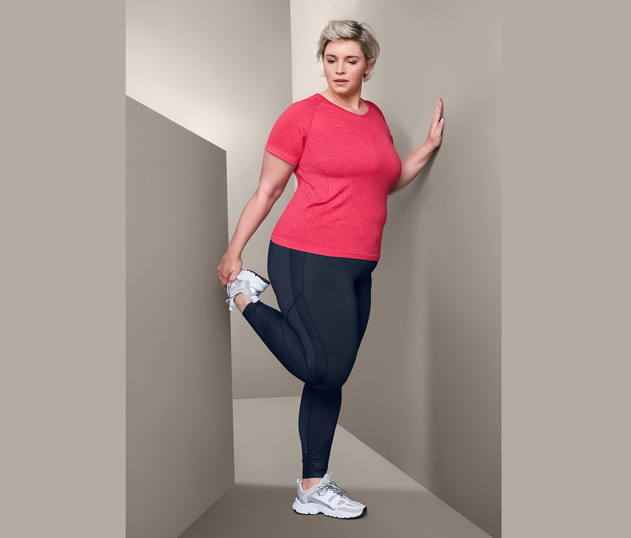 Kadın kırmızı dikişsiz fonksiyonel tişört ve bodyforming spor taytı ile bacağını esnetiyor.