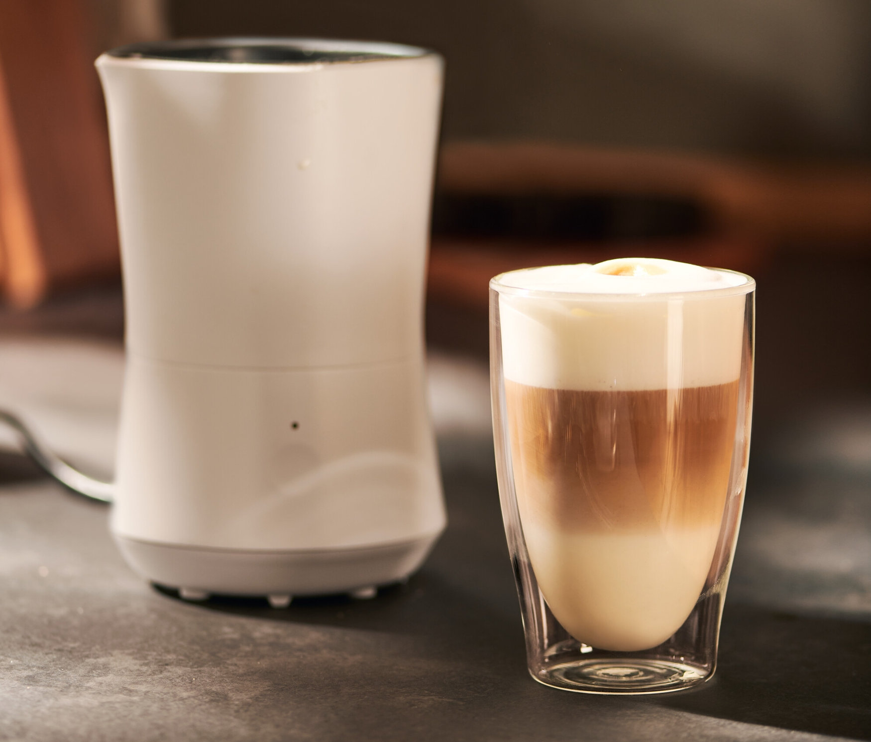 Kremsi beyaz elektrikli süt köpürtücü, üç katmanlı bir içecekle dolu bir bardağın yanında duruyor.