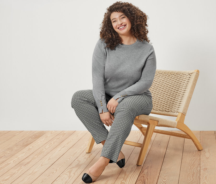 Gülümseyen bir kadın sandalyede oturuyor, üzerinde kırçıllı gri ince örgü kazak ve lacivert baskılı streç pantolon var.