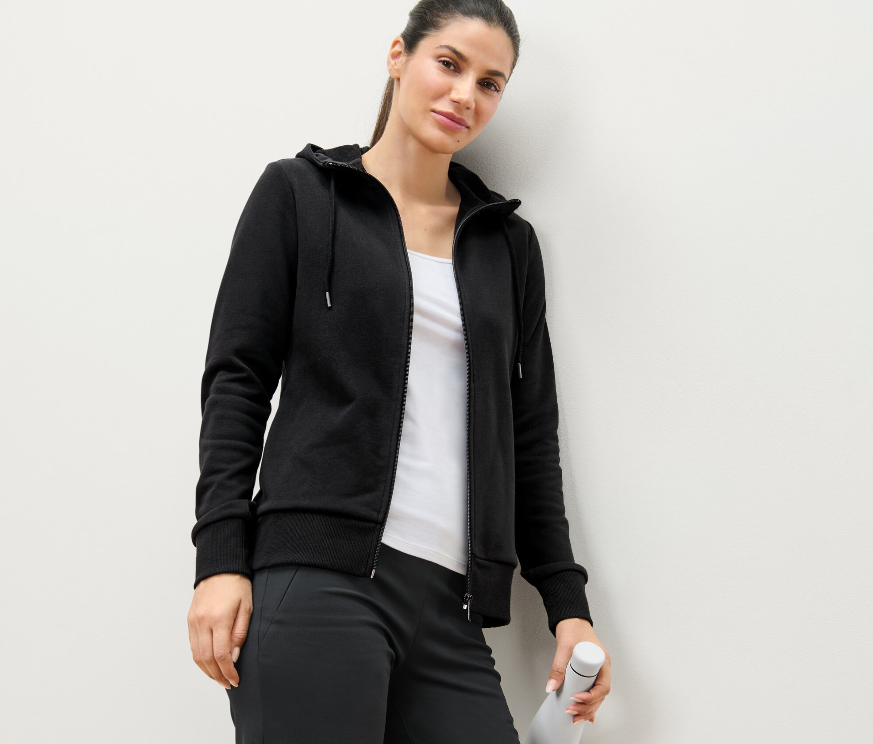 Duvara yaslanmış kadın, siyah sweatshirt mont giyiyor ve beyaz su şişesi tutuyor.