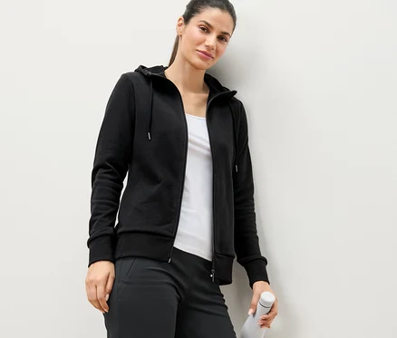 Duvara yaslanmış kadın, siyah sweatshirt mont giyiyor ve beyaz su şişesi tutuyor.