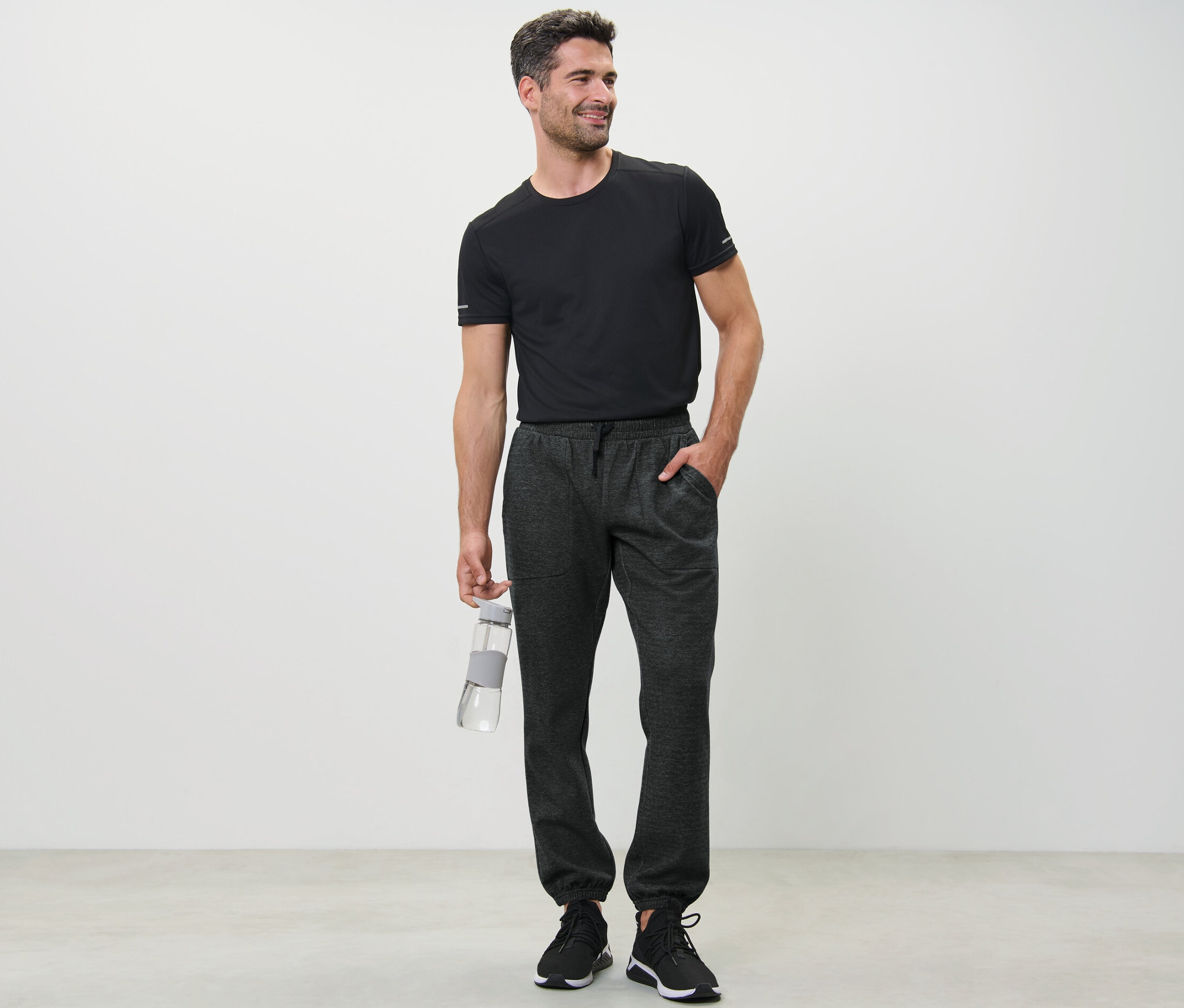 Siyah tişörtlü bir adam su şişesi tutuyor ve eşofman altı giyiyor.