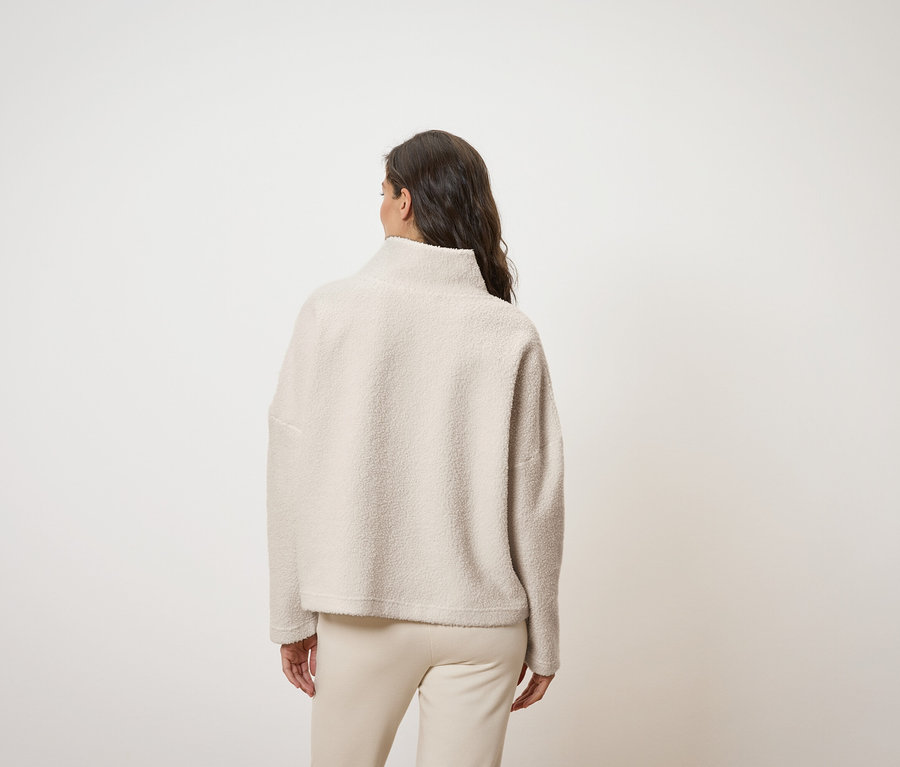 Arkadan gösterilen bir kadın, krem rengi, yüksek yakalı bir üst ve krem rengi pantolon giyiyor.