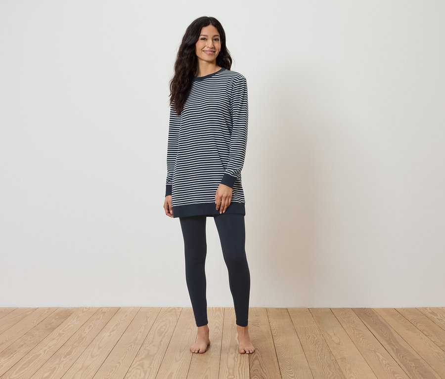 Ahşap zeminde çıplak ayakla duran bir kadın, çizgili uzun kollu bir tişört ve koyu mavi tayt giyiyor.