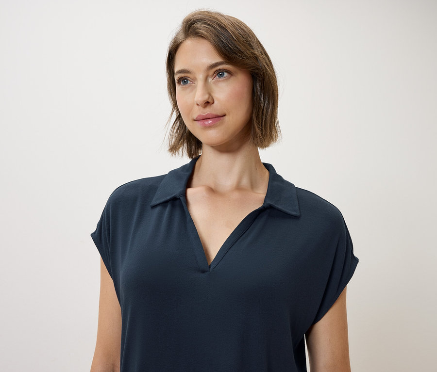 Kahverengi saçlı ve mavi gözlü bir kadın, koyu mavi bir polo elbise giyiyor.