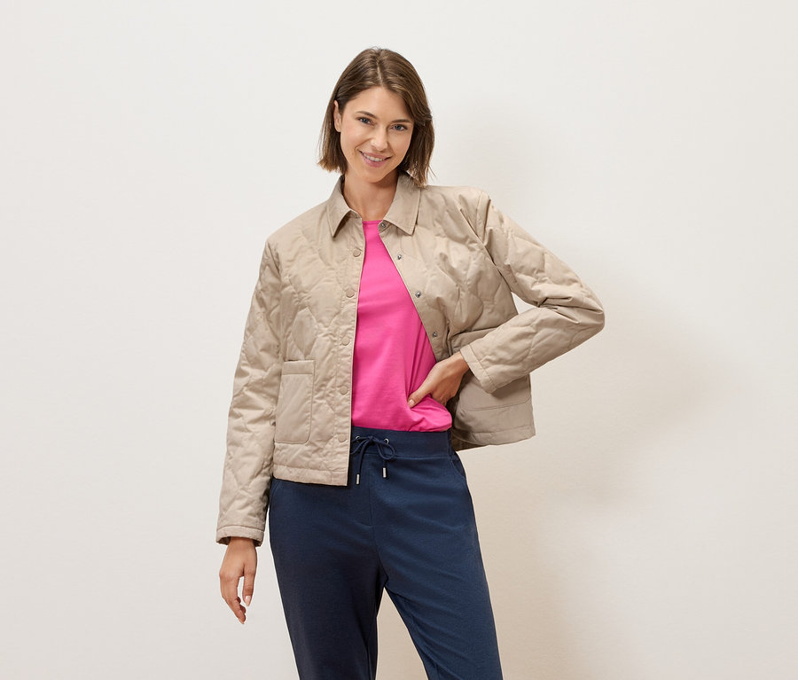 Kısa kahverengi saçlı bir kadın, bej kapitone bir ceket, pembe bir tişört ve mavi pantolonla krem rengi bir duvarın önünde poz veriyor.