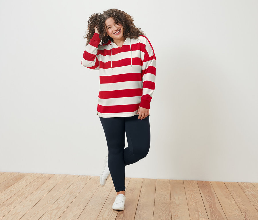 Kıvırcık saçlı, kırmızı beyaz çizgili üst ve mavi taytlı bir kadın ahşap zeminde duruyor.