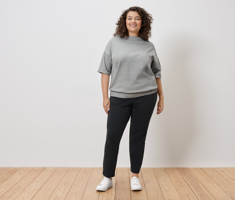 Kıvırcık saçlı bir kadın beyaz bir duvarın önünde duruyor, gri bir sweatshirt, siyah pantolon ve beyaz spor ayakkabılar giyiyor.