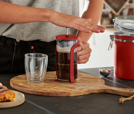 Gözlüklü gülümseyen kadın, ahşap bir tahta üzerindeki kırmızı 300 ml French Press'in pistonunu aşağı bastırıyor, yanında tarçınlı çörekler ve boş bir bardak var.