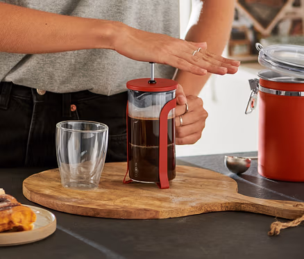 Gözlüklü gülümseyen kadın, ahşap bir tahta üzerindeki kırmızı 300 ml French Press'in pistonunu aşağı bastırıyor, yanında tarçınlı çörekler ve boş bir bardak var.