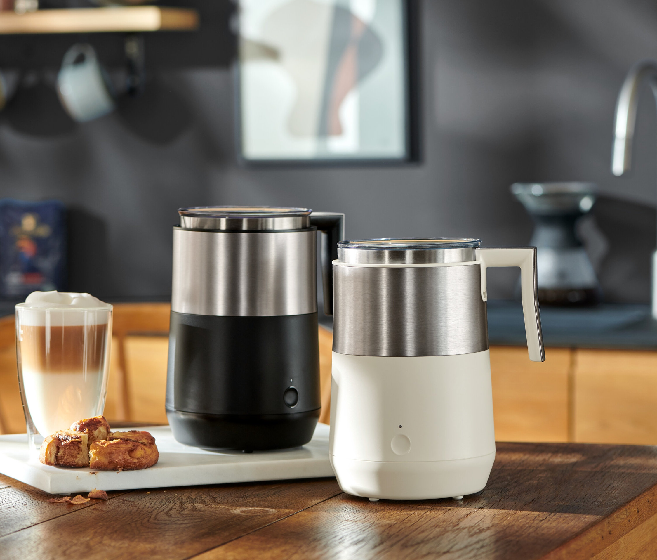 Ahşap bir masa üzerinde bir bardak latte ve hamur işlerinin yanında siyah ve krem rengi olmak üzere iki adet indüksiyonlu süt köpürtücü bulunmaktadır.