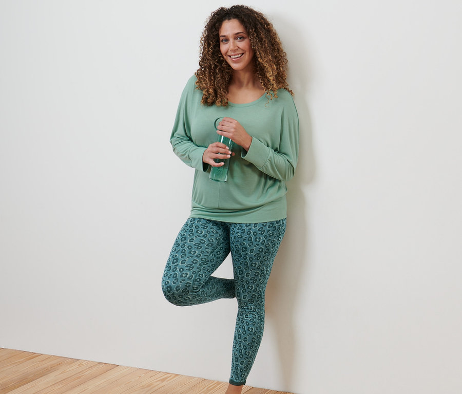 Bir kadın duvara yaslanmış su içiyor. Ada çayı yeşili renginde Spor ve Yoga Üst ve Zümrüt ve Ada Çayı Yeşili renginde Çift Taraflı Spor Taytı giyiyor.