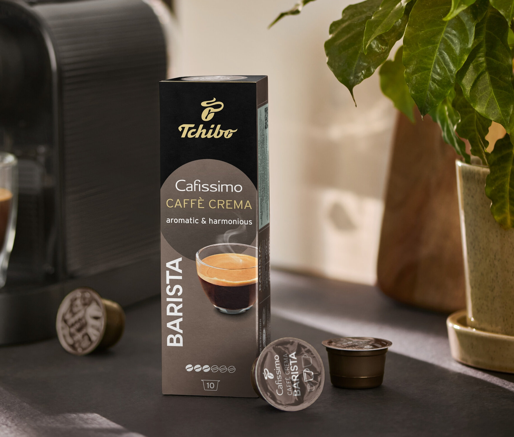 Kahve makinesi bir bardağı kahveyle dolduruyor. Yanında bir paket Cafissimo BARISTA Caffè Crema ve birkaç kapsül duruyor.