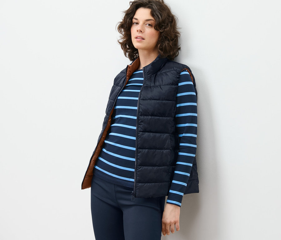 Duvara yaslanmış kadın Çift Taraflı Yelek, çizgili tişört ve pantolon giyiyor.