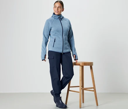 Bir kadın ahşap taburenin yanında duruyor, mavi kapüşonlu sweatshirt ve Softshell Jogging Pantolon giyiyor.