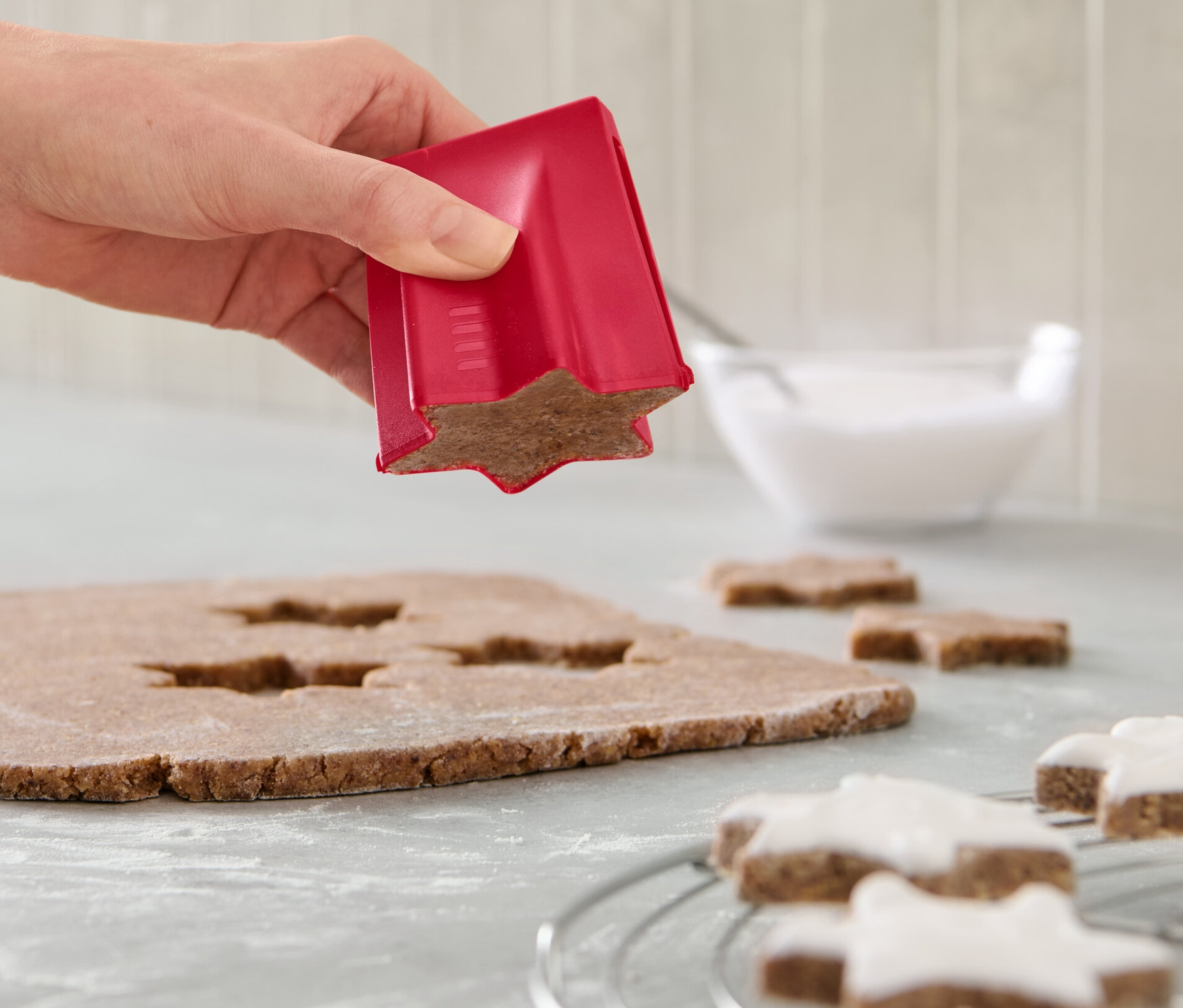 Bir el, hamur üzerinde yıldız kurabiye kalıbı kullanıyor. Tezgahta süslenmiş yıldızlar ve bir kase krema var.