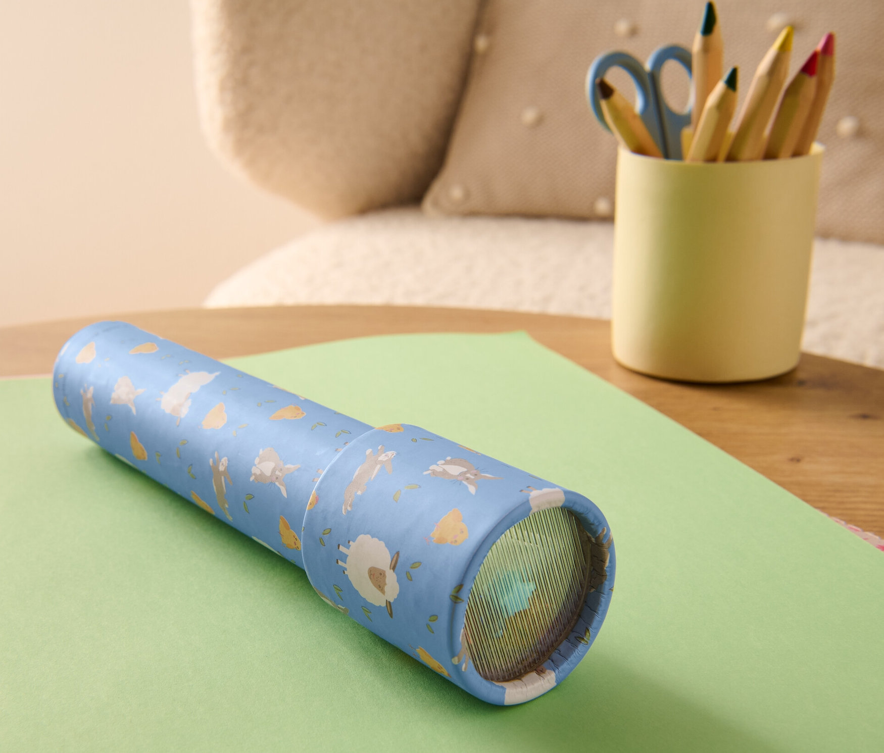 Bir Kaleydoskop yeşil bir kağıdın üzerinde duruyor. Yanında renkli kalemler ve makas olan bir bardak var.