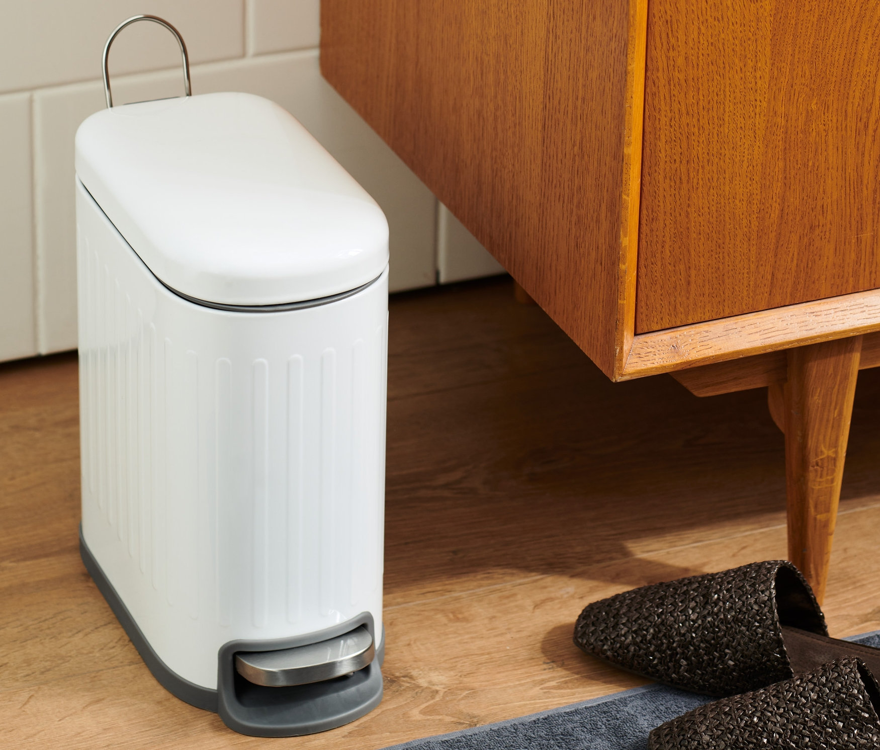 Banyoda gri halı üzerinde kahverengi ahşap dolap ve terliklerin yanında beyaz bir banyo çöp kovası duruyor.